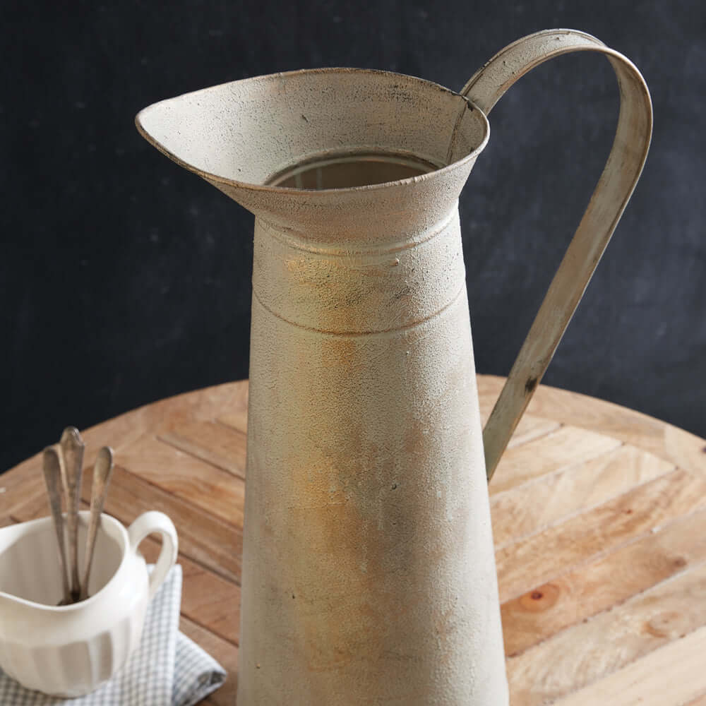 Gilded Farmhouse Tall Pitcher with weathered texture and elegant handle on a wooden table next to a white cup with spoons.